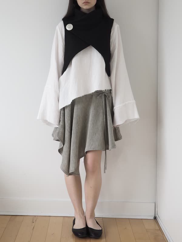 Black crossed cowl vest fastened with a single white button over a white cotton gauze top with wide bell sleeves and raw edges, above an olive wrap skirt with a handkerchief hem.